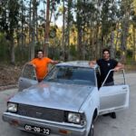 Dois homens posam junto a um automóvel antigo de cor clara, com as portas dianteiras abertas, numa estrada ladeada por árvores. A matrícula visível é GD-09-32.
