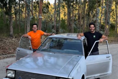 Dois homens posam junto a um automóvel antigo de cor clara, com as portas dianteiras abertas, numa estrada ladeada por árvores. A matrícula visível é GD-09-32.