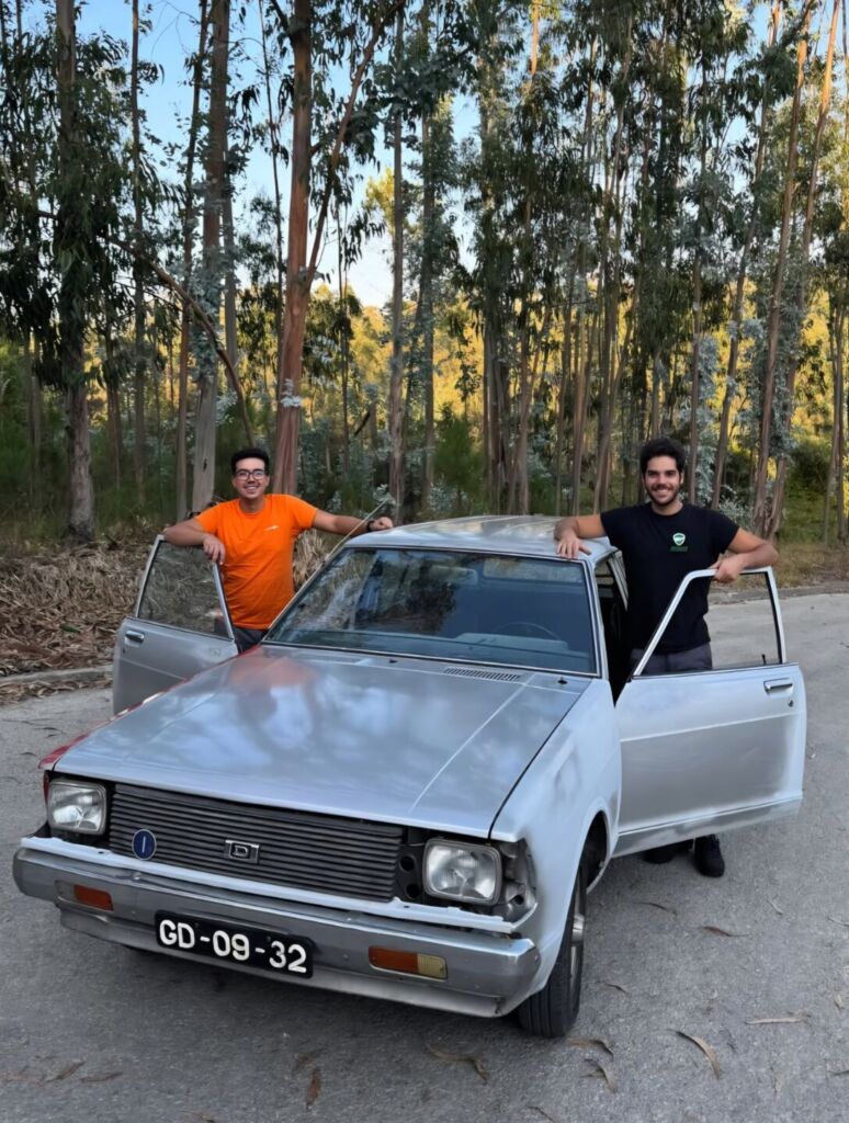 Dois homens posam junto a um automóvel antigo de cor clara, com as portas dianteiras abertas, numa estrada ladeada por árvores. A matrícula visível é GD-09-32.