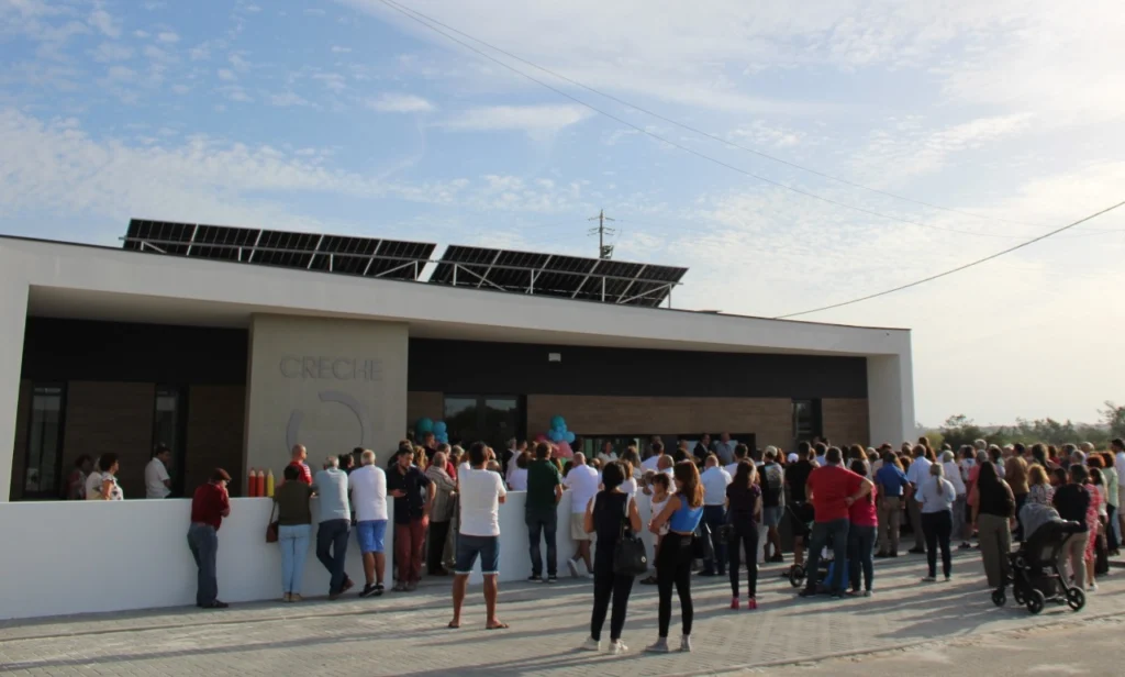 Um grupo de pessoas reúne-se à entrada de um edifício identificado como «Creche». Veem-se painéis solares no telhado e um carrinho de bebé entre os presentes.