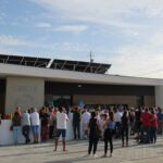 Um grupo de pessoas reúne-se à entrada de um edifício identificado como «Creche». Veem-se painéis solares no telhado e um carrinho de bebé entre os presentes.