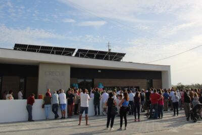 Um grupo de pessoas reúne-se à entrada de um edifício identificado como «Creche». Veem-se painéis solares no telhado e um carrinho de bebé entre os presentes.