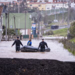 Uma rua está inundada, com a água a atingir as entradas das casas. Duas pessoas com equipamento de proteção deslocam um barco insuflável, junto ao qual está uma terceira pessoa.