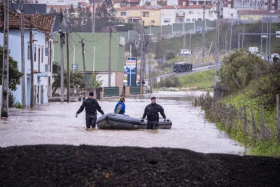 Uma rua está inundada, com a água a atingir as entradas das casas. Duas pessoas com equipamento de proteção deslocam um barco insuflável, junto ao qual está uma terceira pessoa.