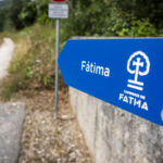 Uma placa azul dos Caminhos de Fátima aponta para um caminho de terra. A placa está junto a uma zona arborizada.