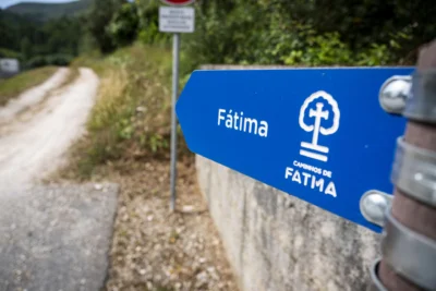 Uma placa azul dos Caminhos de Fátima aponta para um caminho de terra. A placa está junto a uma zona arborizada.