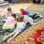 Cinco crianças brincam juntas sobre tapetes coloridos, num espaço ao ar livre. Há fardos de palha e adultos parcialmente visíveis ao fundo.
