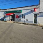 Fachada de um supermercado Meu Super, com toldo vermelho e letreiros visíveis. À frente, vê-se uma rua e um automóvel estacionado; não há pessoas na imagem.