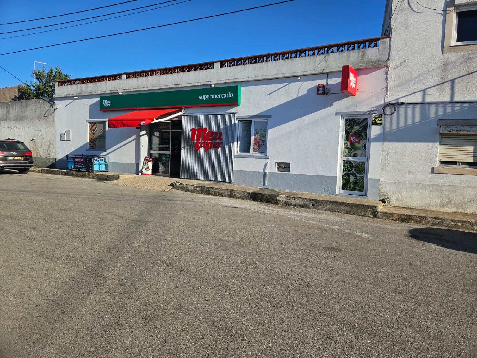 Fachada de um supermercado Meu Super, com toldo vermelho e letreiros visíveis. À frente, vê-se uma rua e um automóvel estacionado; não há pessoas na imagem.