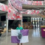 Uma pessoa segura uma bandeira com o brasão e a inscrição «Vila de Ansião» no interior de um edifício. Ao fundo veem-se mesas, cadeiras e uma instalação suspensa com imagens aéreas.