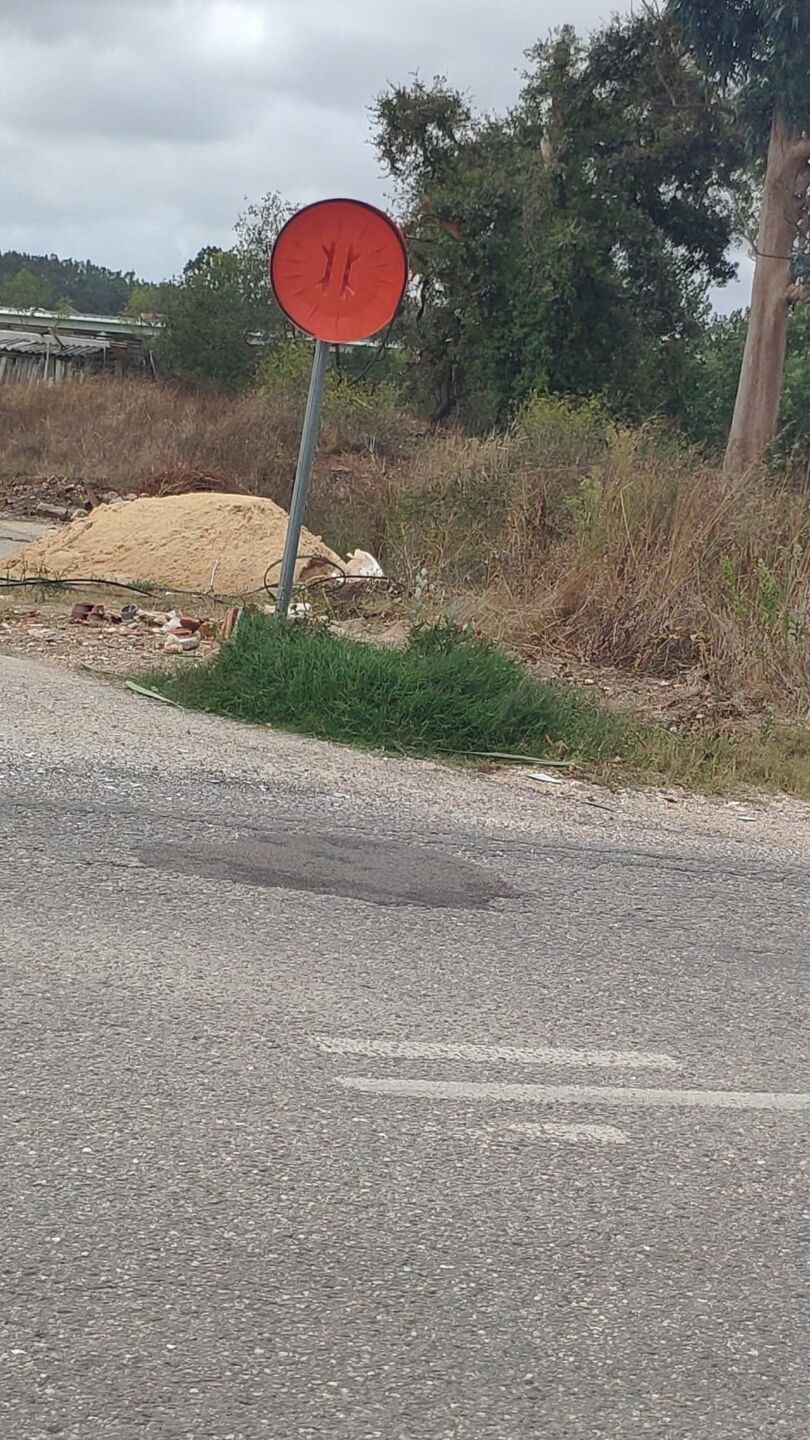 Uma placa circular vermelha, com a inscrição pouco legível, está inclinada junto à berma de uma estrada. Ao lado veem-se um monte de areia, alguns detritos e vegetação seca.
