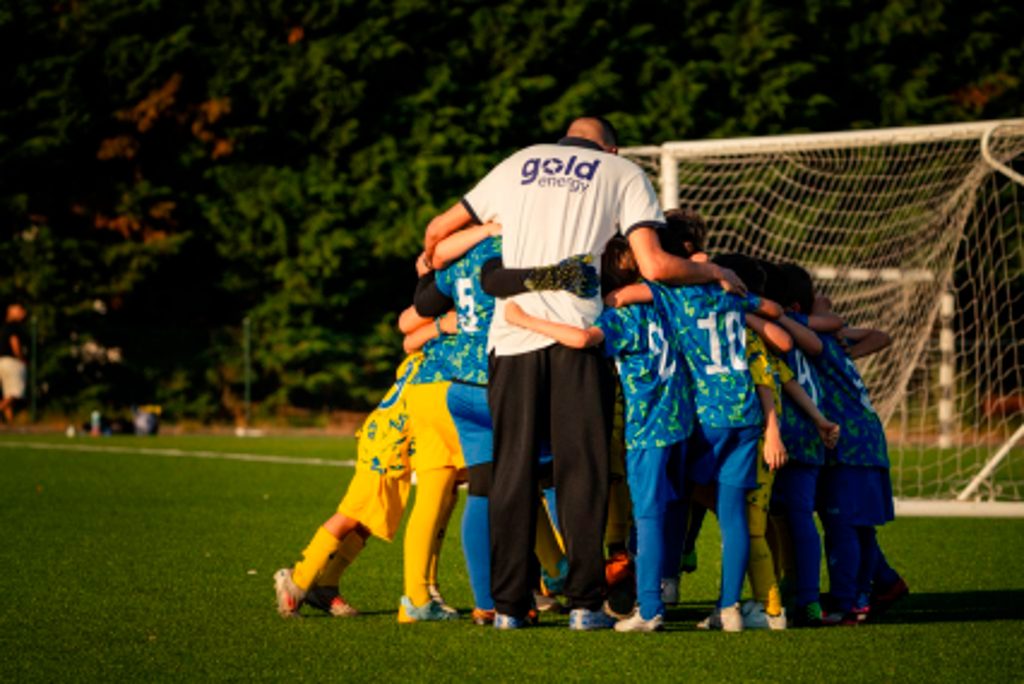 Uma equipa de futebol reúne-se num abraço coletivo junto à baliza, num campo relvado. Os jogadores vestem equipamentos azuis e amarelos.