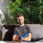 Um homem está sentado num sofá a ler um livro, junto a uma mesa. Há plantas atrás dele e luz solar a entrar no espaço.