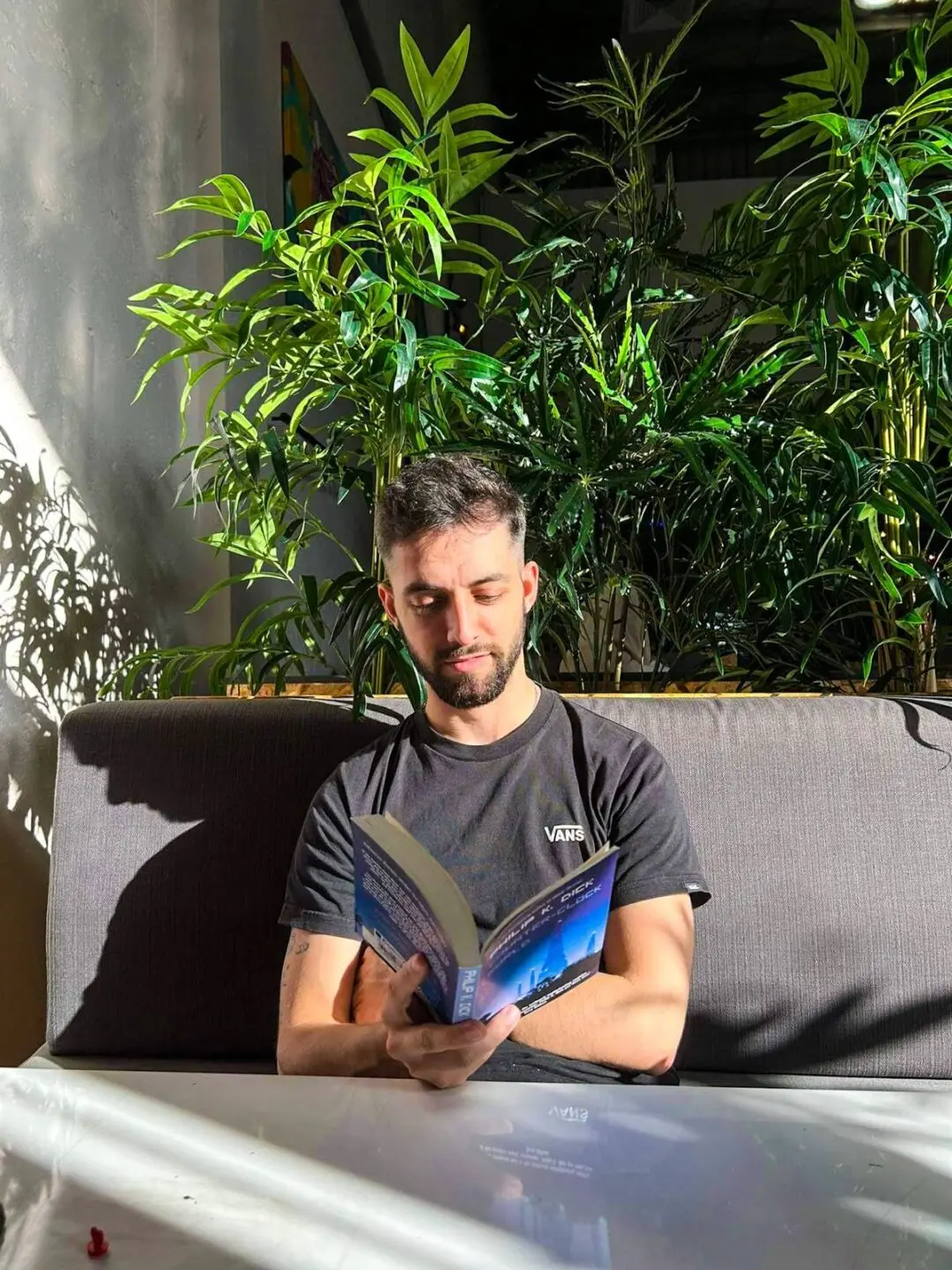 Um homem está sentado num sofá a ler um livro, junto a uma mesa. Há plantas atrás dele e luz solar a entrar no espaço.