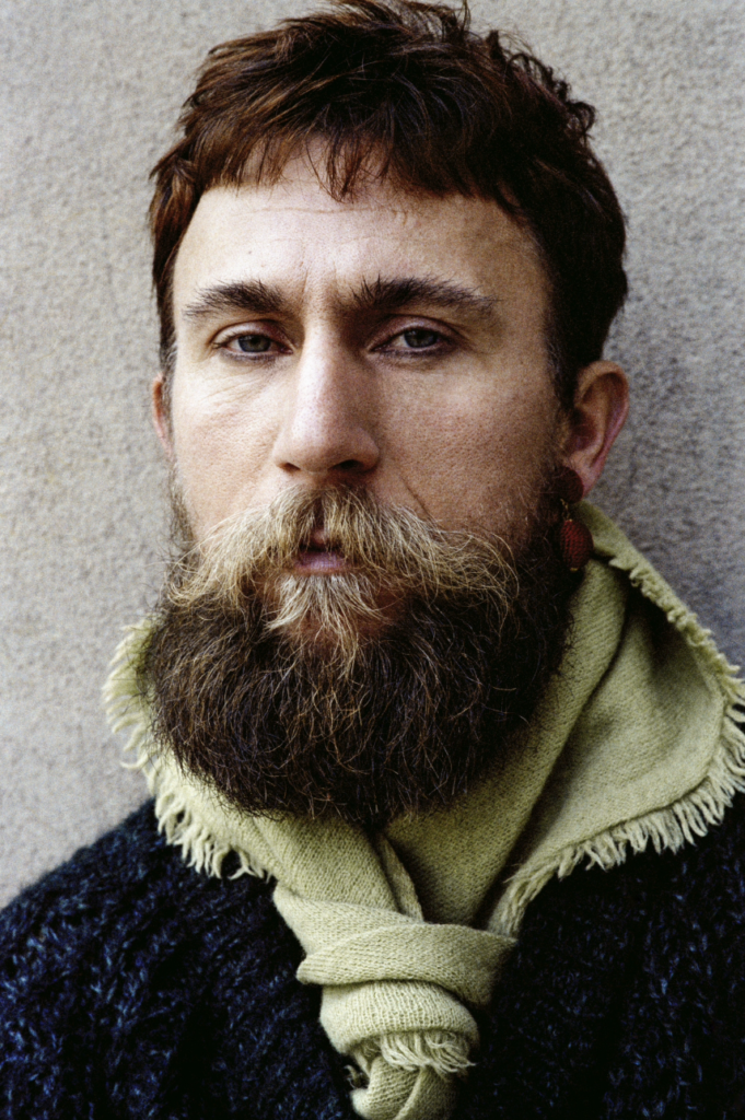 Retrato em grande plano de uma pessoa com cabelo curto e barba comprida, a olhar para a câmara. Usa um cachecol claro e um casaco escuro, diante de um fundo neutro.
