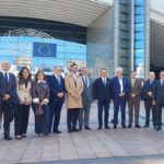 Um grupo de pessoas posa para uma fotografia à entrada de um edifício onde estão visíveis o emblema do Parlamento Europeu e a bandeira da União Europeia.