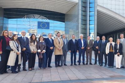 Um grupo de pessoas posa para uma fotografia à entrada de um edifício onde estão visíveis o emblema do Parlamento Europeu e a bandeira da União Europeia.