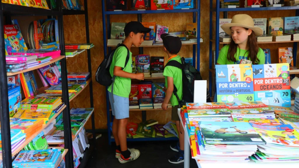 Três crianças observam livros numa livraria ou banca de livros. As estantes e a mesa exibem sobretudo livros infantis e juvenis em português.