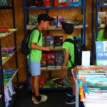 Três crianças observam livros numa livraria ou banca de livros. As estantes e a mesa exibem sobretudo livros infantis e juvenis em português.