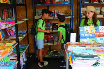 Três crianças observam livros numa livraria ou banca de livros. As estantes e a mesa exibem sobretudo livros infantis e juvenis em português.