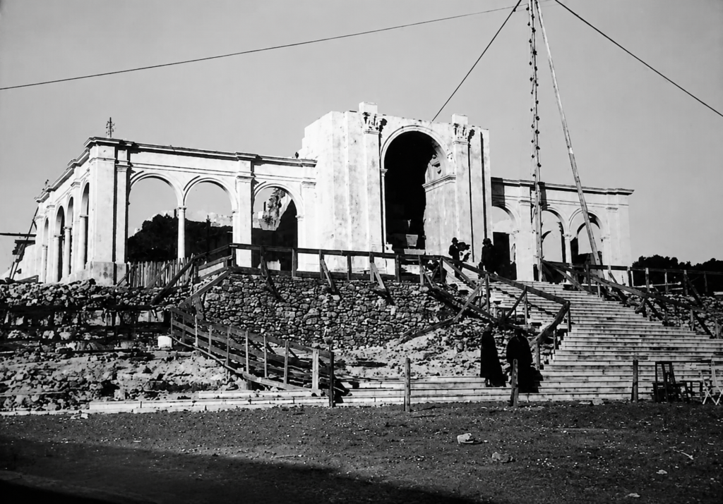 Fotografia a preto e branco de um edifício com arcadas e uma escadaria em construção. Veem-se andaimes, materiais de obra e várias pessoas.