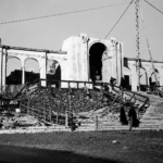 Fotografia a preto e branco de um edifício com arcadas e uma escadaria em construção. Veem-se andaimes, materiais de obra e várias pessoas.