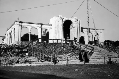Fotografia a preto e branco de um edifício com arcadas e uma escadaria em construção. Veem-se andaimes, materiais de obra e várias pessoas.
