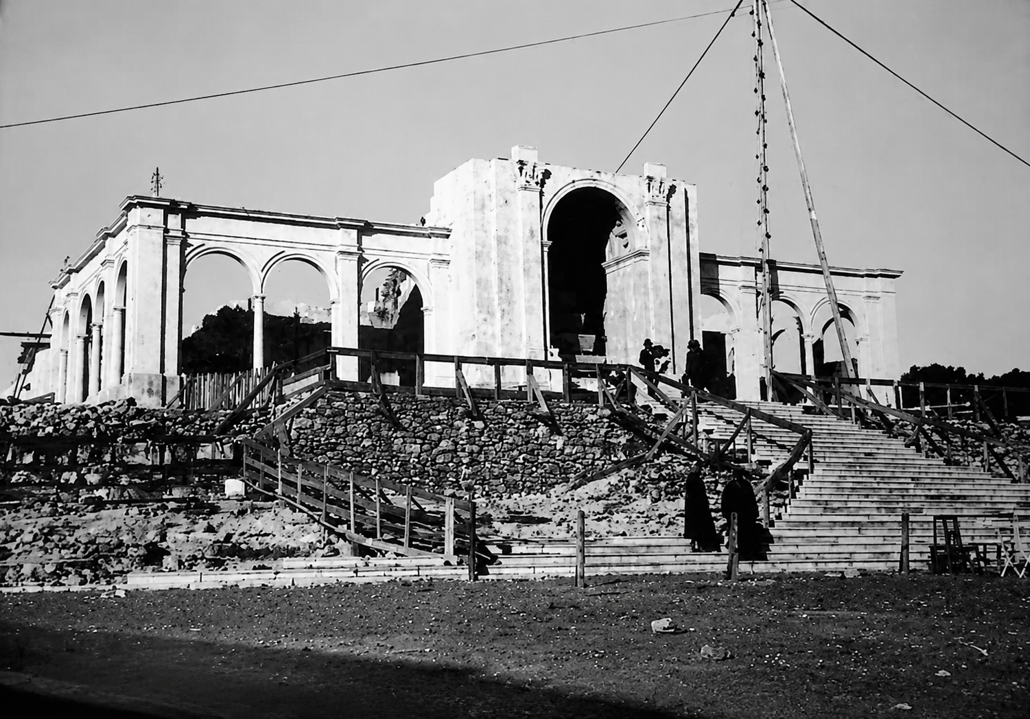Fotografia a preto e branco de um edifício com arcadas e uma escadaria em construção. Veem-se andaimes, materiais de obra e várias pessoas.