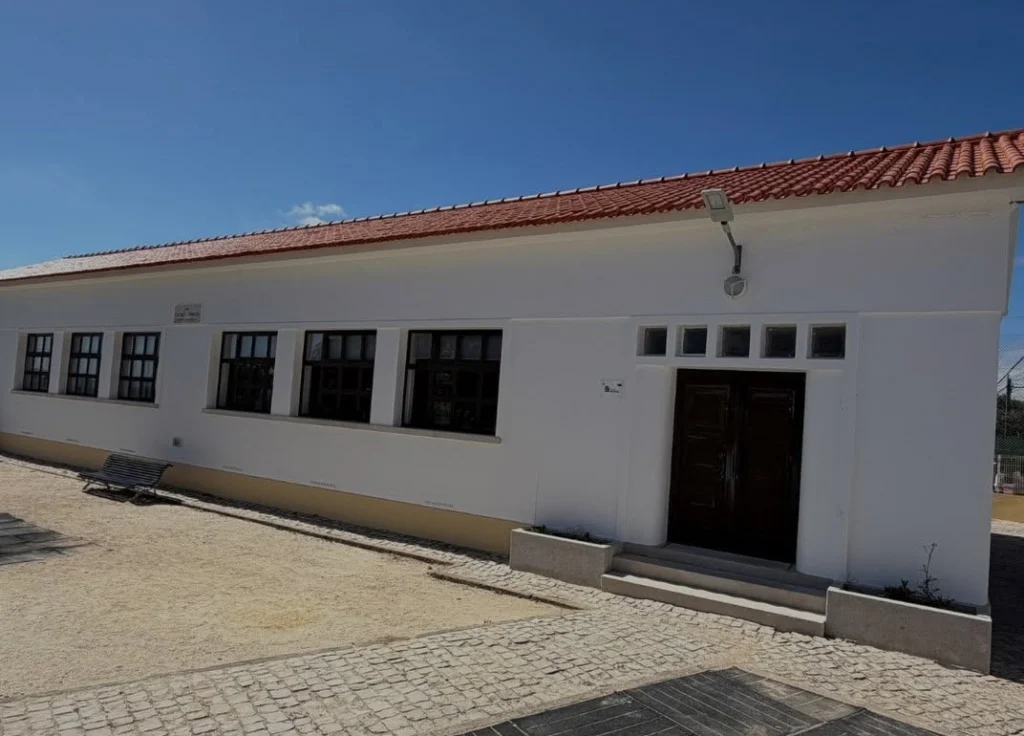 Edifício de um piso, com fachada branca, telhado de telha vermelha e porta de madeira. Em frente, há um banco e uma área pavimentada; não se veem pessoas.