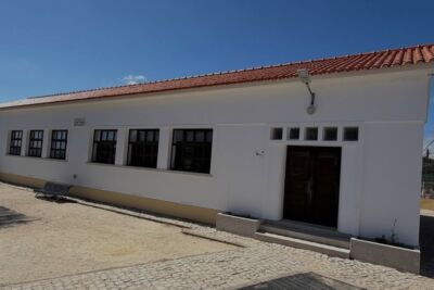 Edifício de um piso, com fachada branca, telhado de telha vermelha e porta de madeira. Em frente, há um banco e uma área pavimentada; não se veem pessoas.