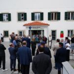Um grupo de pessoas reúne-se à entrada do Centro Social Paroquial de Freixianda. Junto à porta, um homem lê de uma pasta e um sacerdote acompanha a cerimónia.
