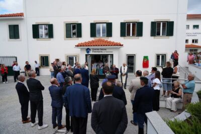 Um grupo de pessoas reúne-se à entrada do Centro Social Paroquial de Freixianda. Junto à porta, um homem lê de uma pasta e um sacerdote acompanha a cerimónia.