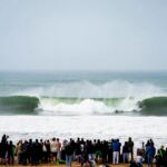 Uma multidão observa da praia dois surfistas em lados opostos de uma grande onda a rebentar. Há câmaras e tripés entre os espectadores.