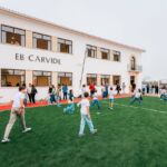 Crianças jogam num campo de futebol junto ao edifício da EB Carvide. Vários adultos acompanham a atividade à volta do campo.