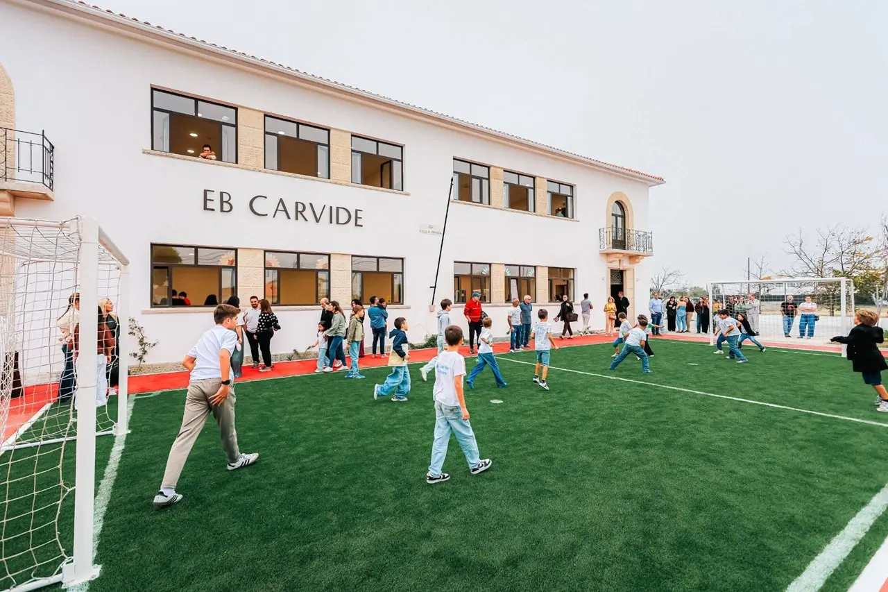 Crianças jogam num campo de futebol junto ao edifício da EB Carvide. Vários adultos acompanham a atividade à volta do campo.