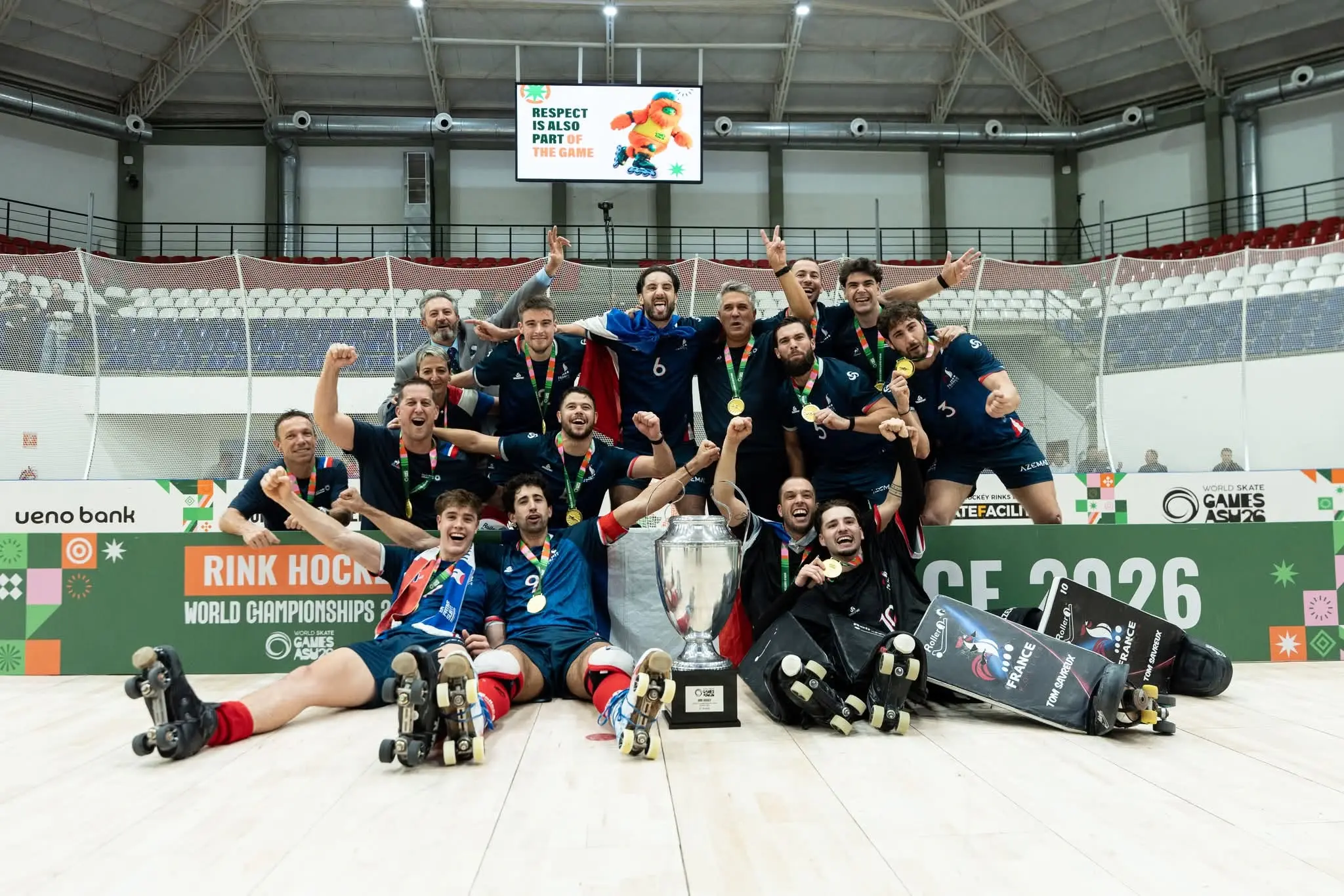 Uma equipa de hóquei em patins posa com medalhas e um troféu num pavilhão. Na imagem são visíveis referências ao Campeonato do Mundo de Hóquei em Patins e aos World Skate Games Asunción 2026.