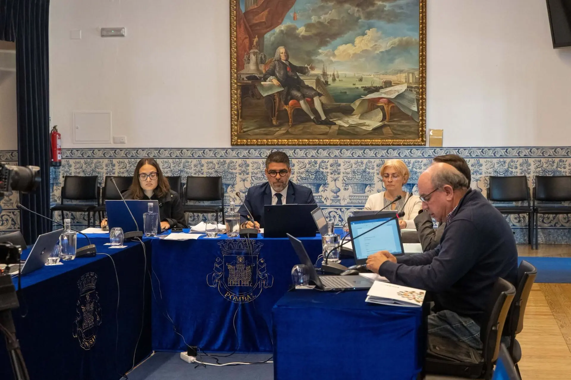 Várias pessoas estão sentadas numa sala de reuniões, com computadores, documentos e microfones. Nas mesas vê-se o brasão e o nome de Pombal.