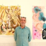 Um homem posa numa galeria diante de duas obras de arte: uma pintura com coelhos e árvores e uma composição abstrata em tons de rosa e azul.