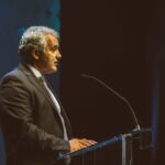 Um homem de fato e gravata fala ao microfone, atrás de um púlpito transparente, num palco. A imagem não contém texto visível que permita identificá-lo ou determinar o evento.