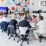 Um grupo de pessoas participa numa reunião à volta de uma mesa, com documentos e um ecrã de apresentação. Na parede lê-se «EPNAZARÉ escola profissional».