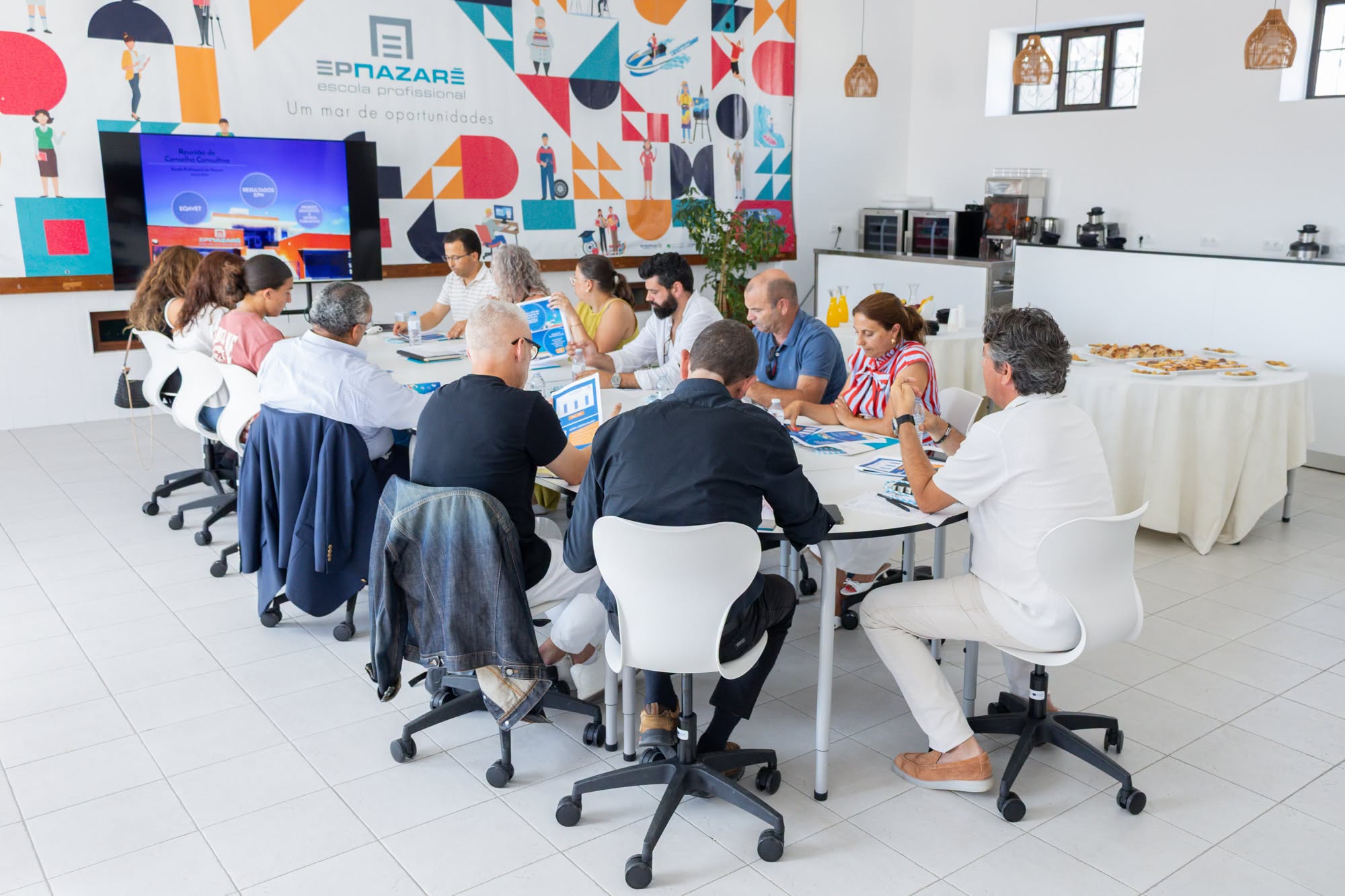 Um grupo de pessoas participa numa reunião à volta de uma mesa, com documentos e um ecrã de apresentação. Na parede lê-se «EPNAZARÉ escola profissional».