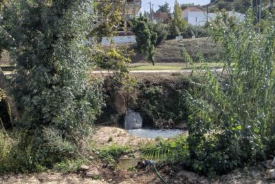 Um pequeno curso de água passa por uma zona com vegetação densa e uma queda de água. Ao fundo, veem-se edifícios, incluindo uma construção em obras.