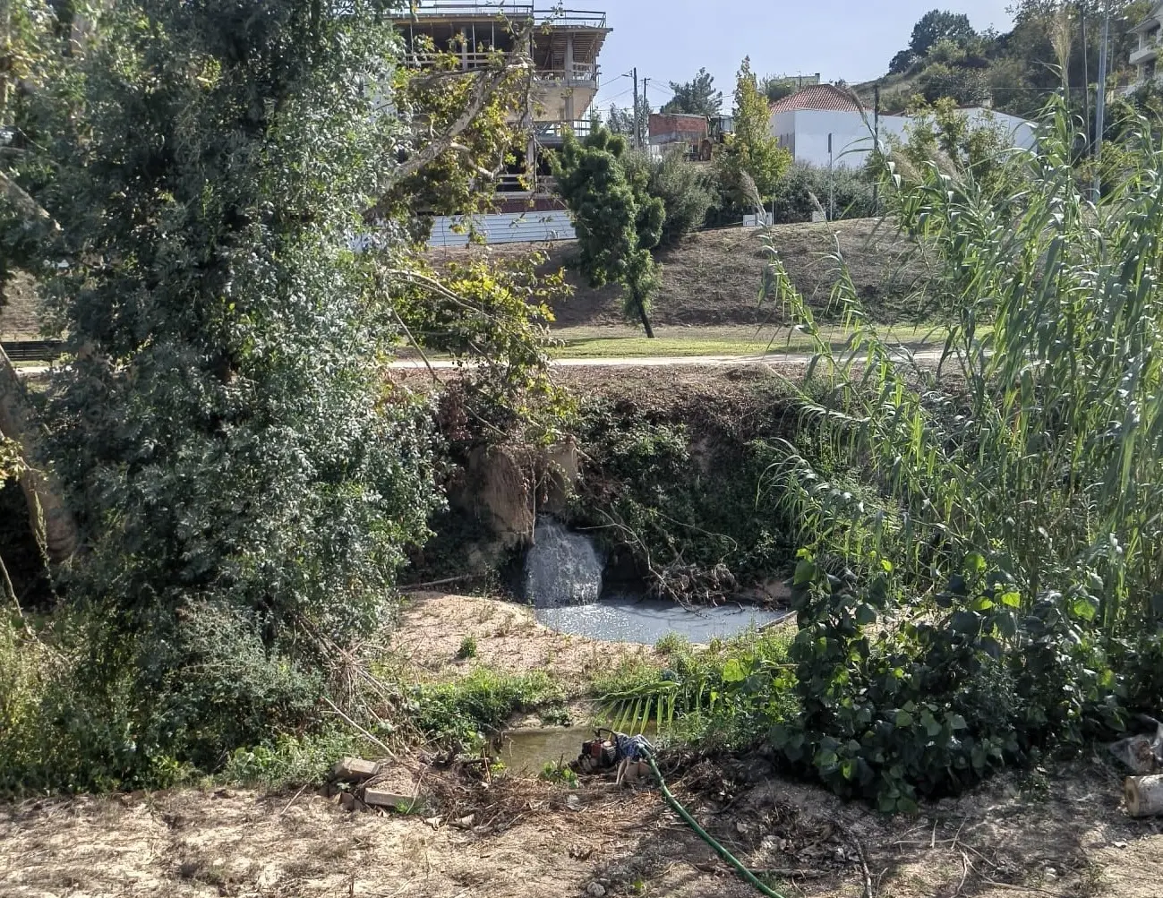 Um pequeno curso de água passa por uma zona com vegetação densa e uma queda de água. Ao fundo, veem-se edifícios, incluindo uma construção em obras.