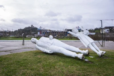 Duas esculturas brancas de figuras humanas, inclinadas e próximas do chão, estão num espaço verde. Ao fundo, veem-se edifícios, estruturas em construção e um castelo no alto de uma colina.