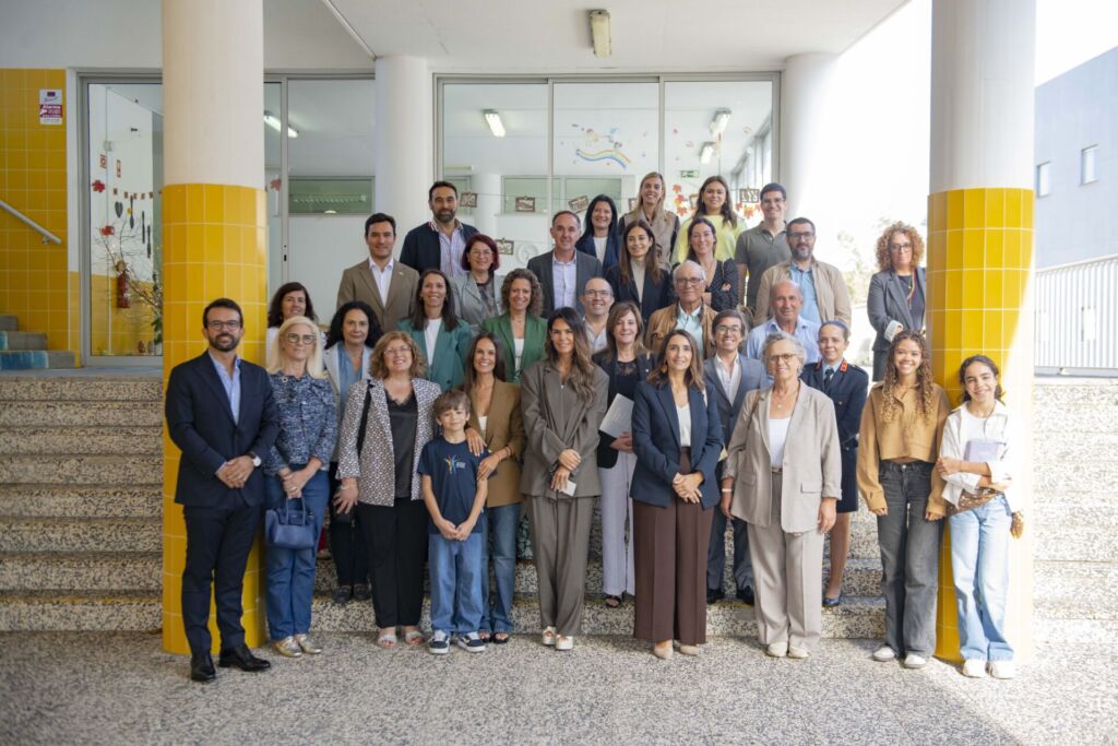 Fotografia de grupo à entrada de um edifício, com adultos, jovens e uma criança dispostos em vários degraus. Veem-se portas de vidro, colunas com azulejos amarelos e decorações coloridas no interior.