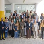 Fotografia de grupo à entrada de um edifício, com adultos, jovens e uma criança dispostos em vários degraus. Veem-se portas de vidro, colunas com azulejos amarelos e decorações coloridas no interior.
