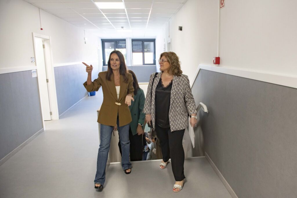 Duas mulheres caminham e conversam num corredor interior, junto a uma escada. Uma delas aponta para a esquerda; outras pessoas seguem atrás.