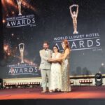 Três pessoas posam em palco com um troféu diante de um painel dos World Luxury Hotel Awards. A imagem sugere um momento de entrega de prémio, sem identificar os participantes ou a distinção atribuída.