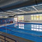 A imagem mostra uma piscina interior vazia, com pistas delimitadas por separadores e bandeirolas suspensas. São visíveis janelas amplas e condutas de ventilação no teto.