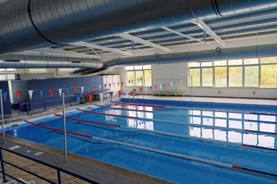 A imagem mostra uma piscina interior vazia, com pistas delimitadas por separadores e bandeirolas suspensas. São visíveis janelas amplas e condutas de ventilação no teto.